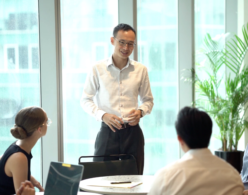 Happy people sat around an office table listening to a standing man who is smiling through his words